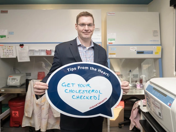 Tall, white man in dark blue sport jacket stands in a lab holding a heart poster. 