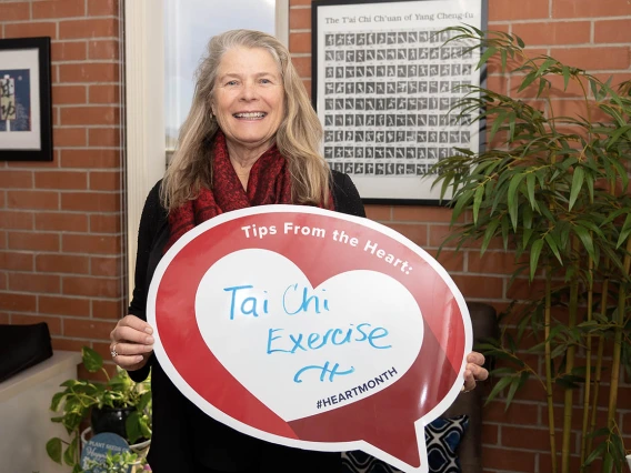 Middle-aged white woman standing in front of brick wall and plant holding heart poster.