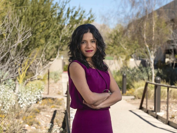 South Asian woman wearing a maroon dress stands outside with her arms crossed and a smiling face