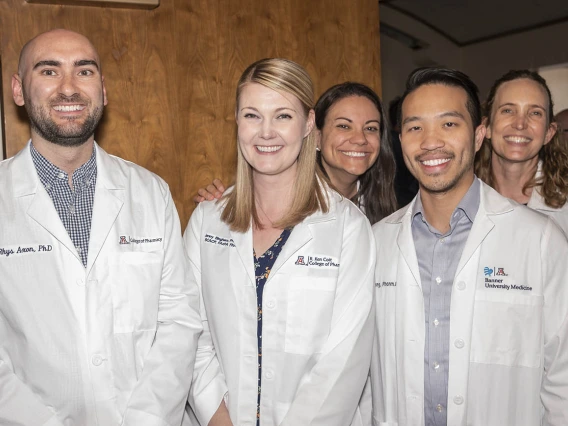 Five men and women Pharmacy faculty members wearing white coats standing side-by-side smiling. 