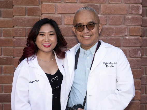 A young Asian woman in a pharmacy white coat stands with her father in a medical white coat, both smiling. 