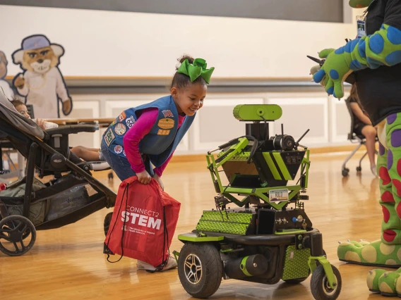 A young girl scout leans over looking closely at a green robot. 