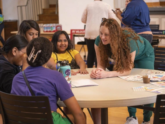 A young female medical student leans over a round table talking with a group of girl scouts.