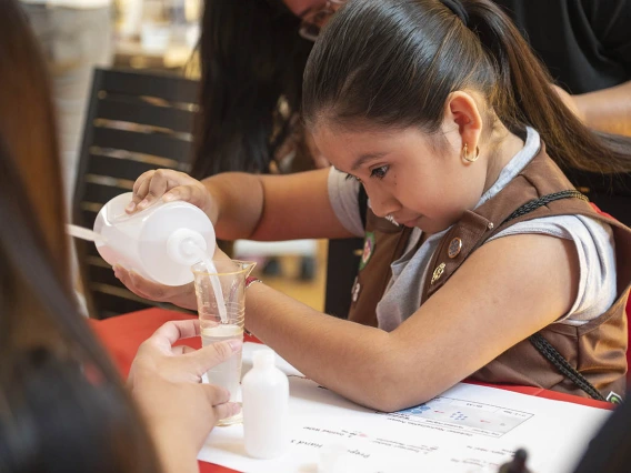 A young girl scout with dark hari in a ponytail pours liquid into a container at an activity table. 