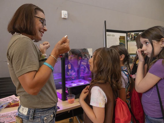 A middle-aged woman with dark brown hair stands next to several girl scouts explaining a display at a table. 