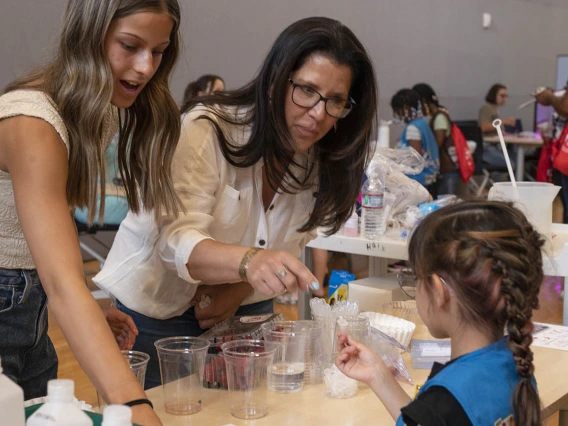 A young woman and her mother stand at an activity table helping a young girl scout.