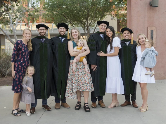 Spouses and children of UArizona College of Medicine – Phoenix class of 2022 graduates pose for a photo after the commencement ceremony.  The graduates, in black gowns from left are, Brendon Warner, MD, Jordan Yergensen, MD, Connor Swensen, MD, and Scott Litton, MD.