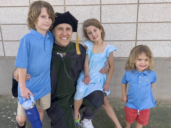 A light-skinned young man in graduation regalia kneels down and is surrounded by his three young children. 