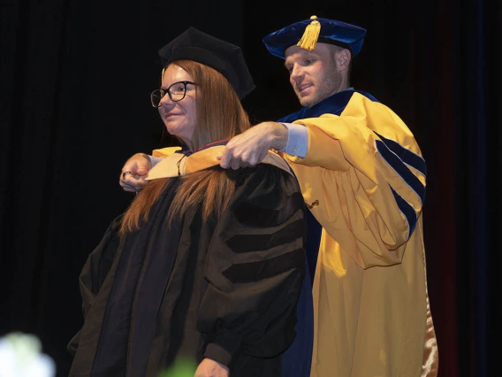 A young woman with red hair and wearing graduation regalia receives her hood by a male professor during convocation.