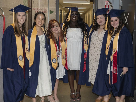 Six female graduates dressed in cap and gown wait backstage in Centennial Hall before receiving their degrees.