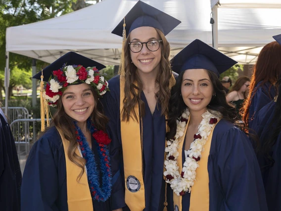 Three smiling women dressed in graduation regalia stand outside after their convocation. 