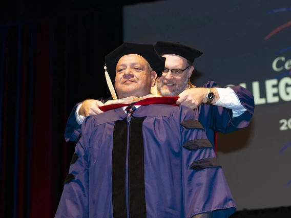 A man dressed in graduation regalia receives his hood from a male professor during graduation.