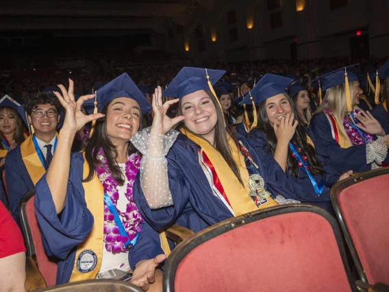 Two young women dressed in graduation regalia flash the University of Arizona hand sign while seated in Centennial Hall.