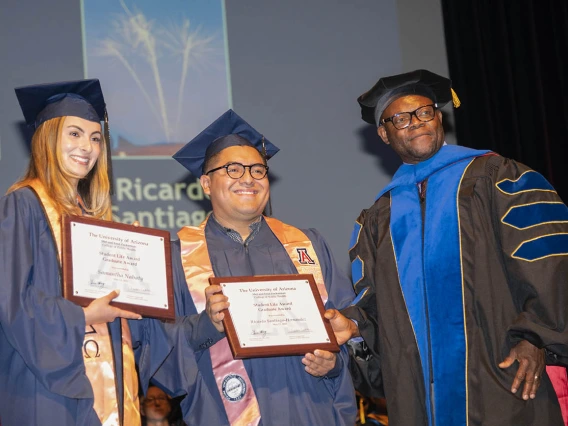 A female and male graduate in caps and gowns stand holding certificates next to a faculty member, also in graduation regalia. 