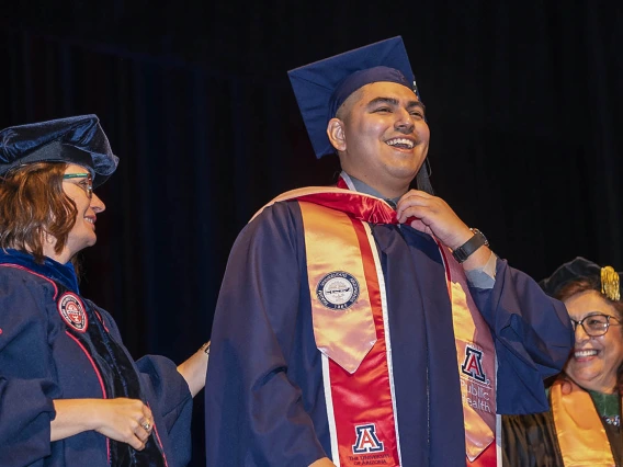 A smiling young man wearing a graduation cap and gown stands in front of two femal professors, also in graduation regalia. 