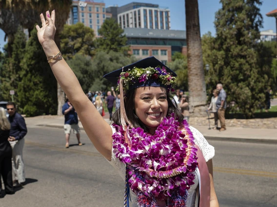 A smiling young woman wearing a graduation cap with several flower garlands around her neck waves while standing outside. 