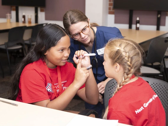 A young nursing student in blue scrubs watches as two girls check each other's pupils with a flashlight.