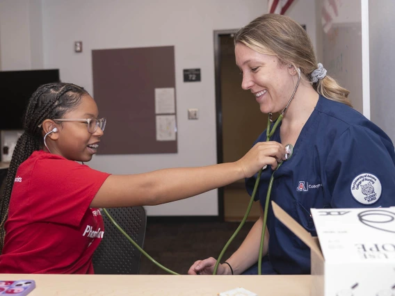 A young dark-skinned girl uses a stethoscope to listen to the heartbeat of a nursing student. 