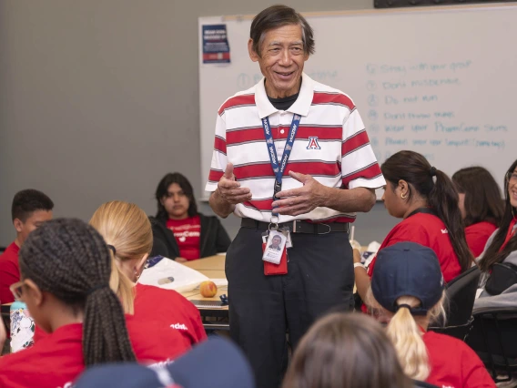 Older Asian man stands in a classroom setting surrounded by kids who are listening to him talk. 