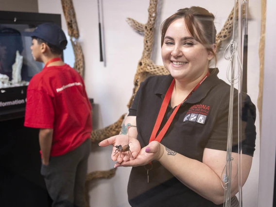 A young light-skinned woman with brown hair smiles as she holds a large bug in her outstretched cupped hands.