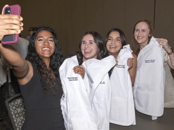 A group of four young women smile as they hold up their medical school white coats as one of them takes a selfie of the group. 