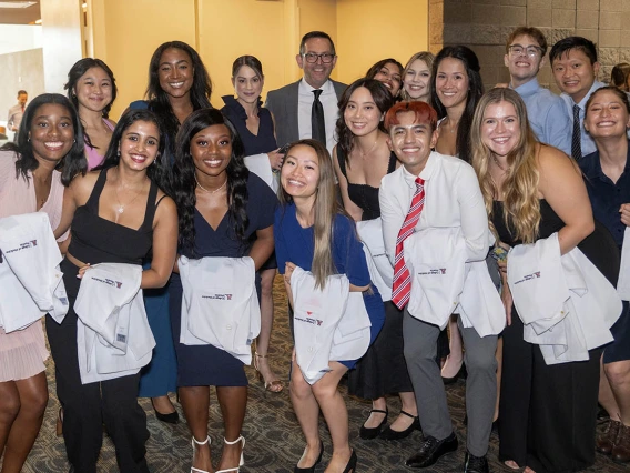 A group of about 16 medical students holding white coats over their arms pose for a photo with a male faculty member. All are smiling. 