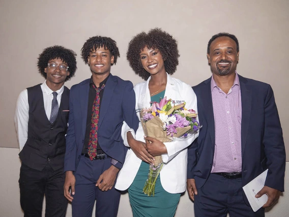 A young female medical student with dark skin smiles holding flowers as she stands withher family.