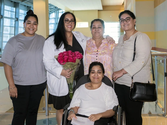 A young woman in a white coat holding flowers and smiling stands with four female members of her family.