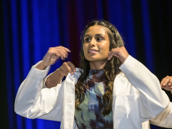 A young, brown-skinned femal medical student with long dark hair looks puts on her white medical coat.