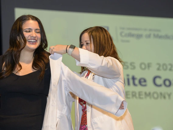 A young female medical student with long dark hair smiles as a female faculty member helps her put on her white coat. 