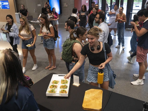 A young woman reaches for a small salad plate from a counter as dozens of other people mingle in the background. 