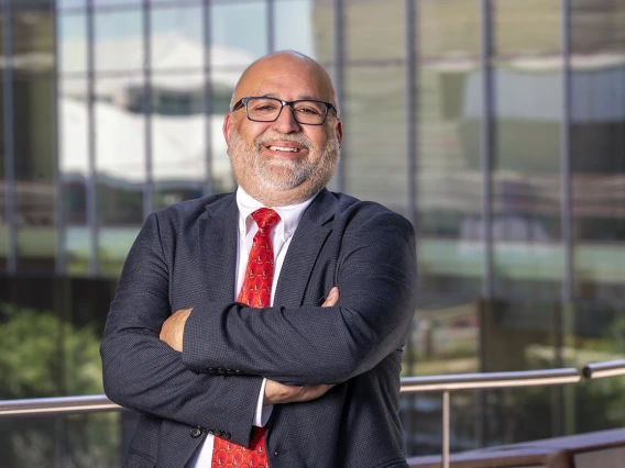 South Asian man with grey beard stands wearing a black suit, white shirt and red tie. His arms are folded and he is smiling.