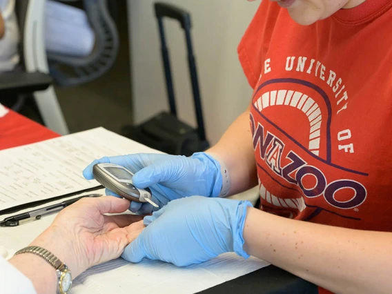 A woman in a red University of Arizona t-shirt wearing medical gloves takes a blood sample from the finger of someone's hand.