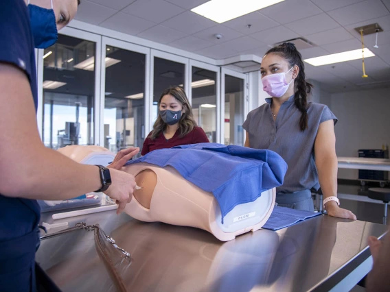 AGACNP student Katie Pon Rambo, in gray scrubs, watches as another student practices inserting a chest tube. This training is required for graduates to apply for credentials post-graduation.  