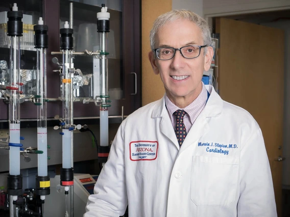 An older white man in a white lab coat stands in a lab smiling. 