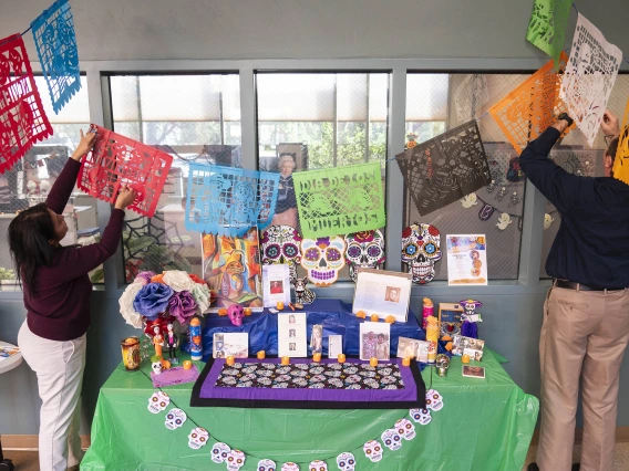 Two people hang tissue paper decorations over a brightly decorated Dia de los Muertos altar with skull decorations and photographs. 