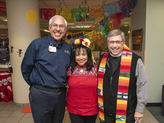 Two men stand on each side of a short woman, all smiling. They are in front of the Dia de los Muertos altar. 