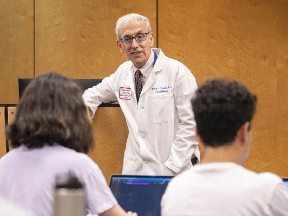 Dr. Marvin Slepian stands in the front of a classroom leaning on the podium while students take notes. 