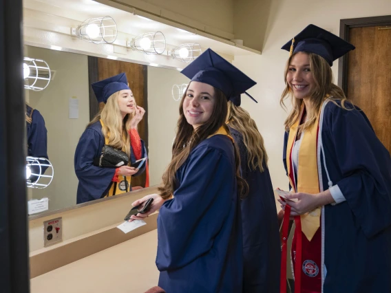 Three young women in graduation caps and gowns stand in a dressing room with a mirror. Two are smiling and one is putting lip gloss on in the mirror.