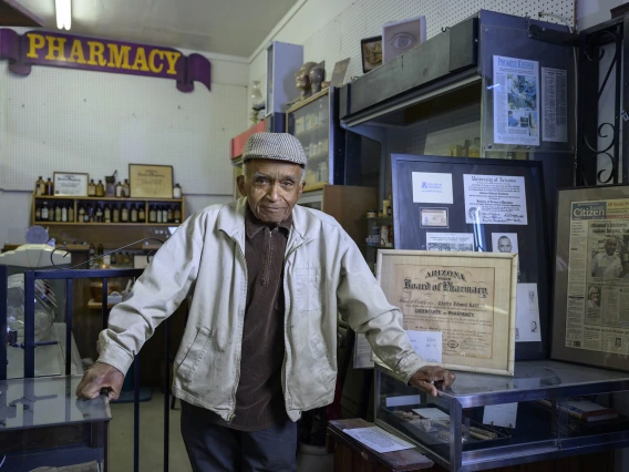 Charles Kendrick stands beneath a sign reading pharmacy and is surrounded with other pharmacy memorabilia.