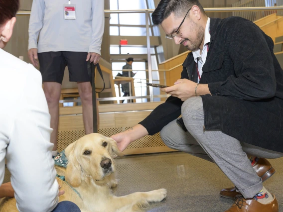 A man in a sport coat squats down to pet a golden retriever. 