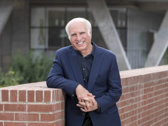 Older man with white hair wearing a navy blue suit leans against a half-brick wall outside smiling. 