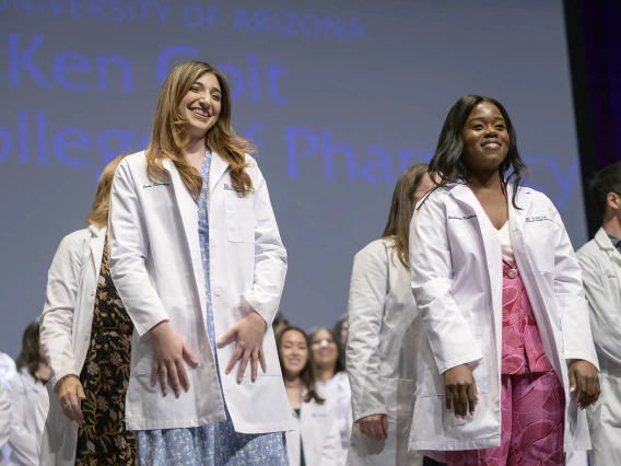 Two University of Arizona College of Pharmacy students smile as they stand in front of other students, all wearing their white coats. 