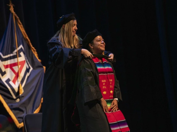 A University of Arizona College of Medicine – Phoenix student in graduation regalia smiles as a professor places a graduation hood on her.