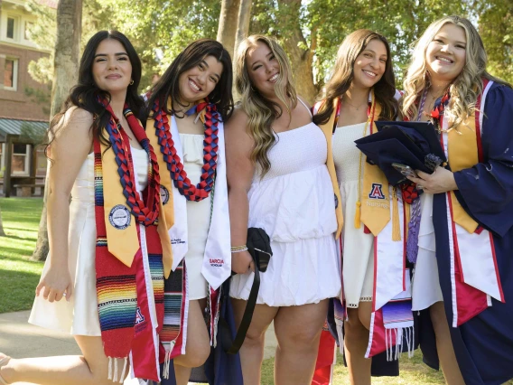 Five University of Arizona College of Nursing Bachelor of Science in Nursing students smile as they stand together outside. Some are wearing graduation gowns and have University of Arizona sashes on. 