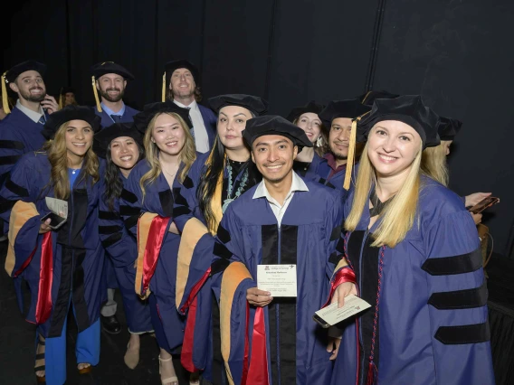 About a dozen smiling University of Arizona College of Nursing students wearing graduation caps and gowns stand backstage waiting for their convocation ceremony to begin. 