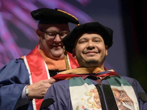 A University of Arizona College of Nursing professor places a hood over the shoulders of a nursing student. Both are dressed in graduation caps and gowns. 