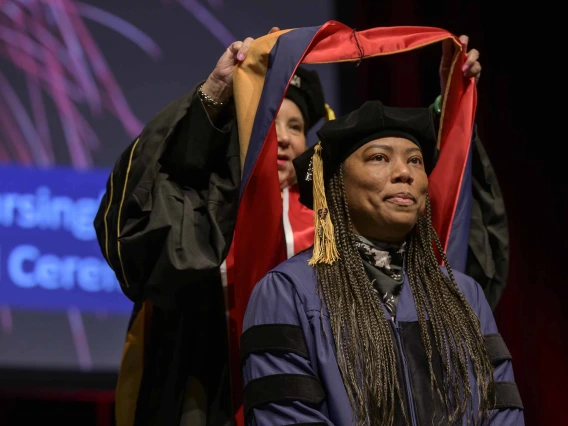 A University of Arizona College of Nursing professor places a hood over the shoulders of a nursing student. Both are dressed in graduation caps and gowns.