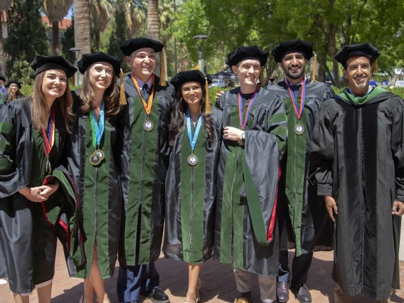 Six University of Arizona College of Medicine – Tucson students and one professor, all in graduation regalia, stand together outside before going to their commencement ceremony. 