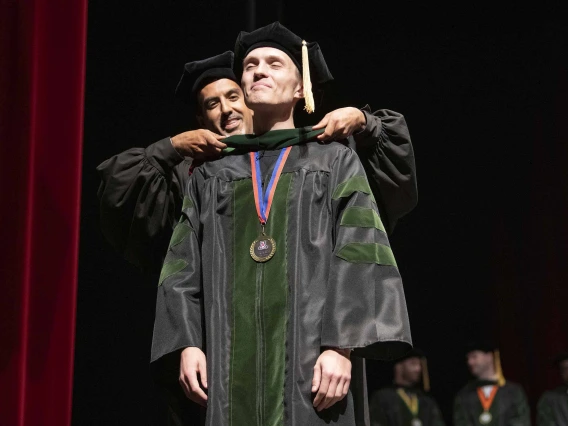 A University of Arizona College of Medicine – Tucson student dressed in a graduation cap and gown has a ceremonial hood placed over his shoulders by his faculty mentor, who is also dressed in graduation regalia.